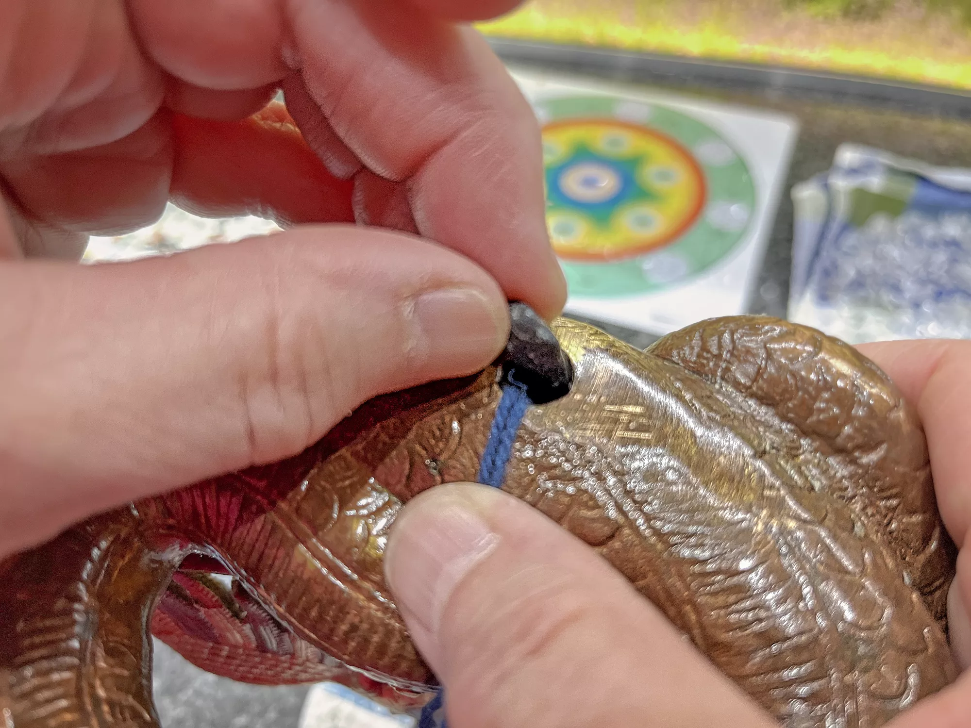 Hände fädeln eine blaue Schnur durch ein geschnitztes, schlangenförmiges Metallobjekt; im Hintergrund liegen farbige Dekorstücke und Verpackungsmaterial.