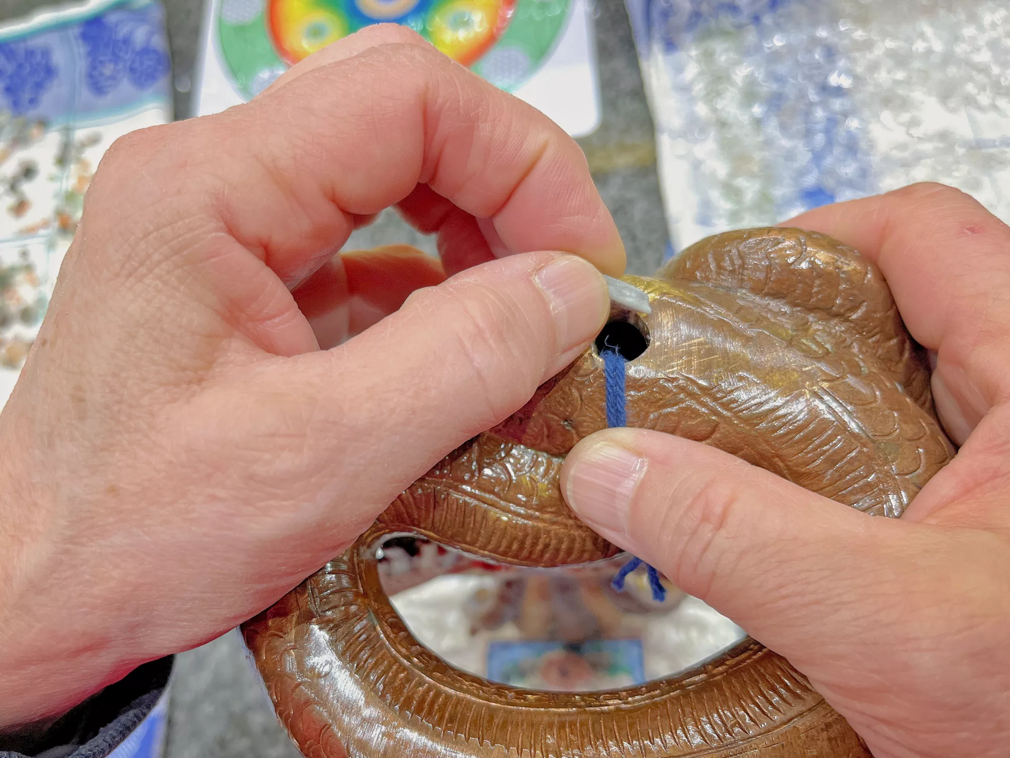 Hände fädeln eine blaue Schnur durch ein geschnitztes, schlangenförmiges Metallobjekt; im Hintergrund liegen farbige Dekorstücke und Verpackungsmaterial.