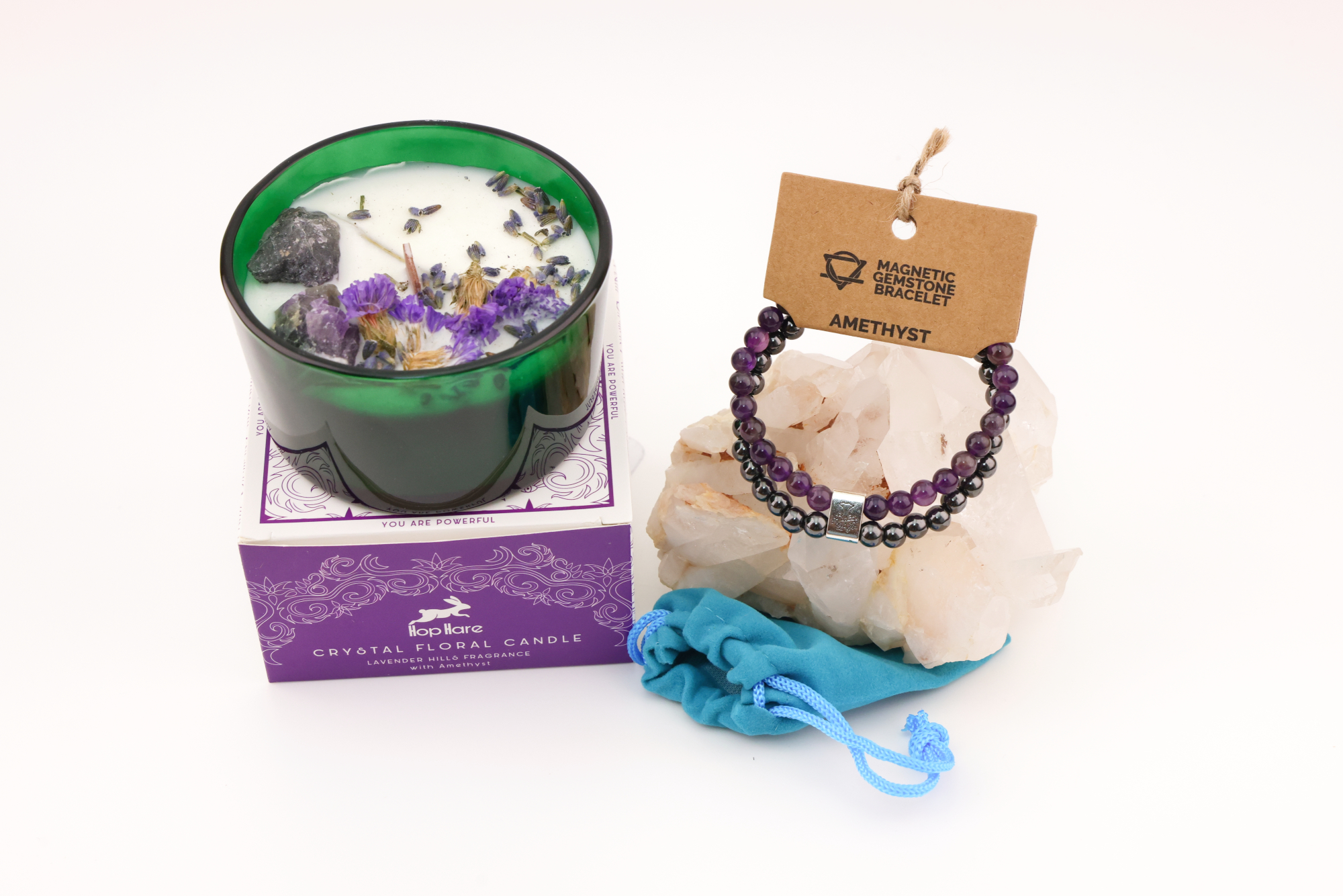 Grüne Hop‑Hare Duftkerze mit Citrin‑ und Amethyst‑Steinen sowie Blüten, daneben die lilafarbene Produktbox; rechts ein Amethyst‑Magnetarmband auf brauner Karte, präsentiert auf Bergkristallen mit türkisfarbenem Beutel.