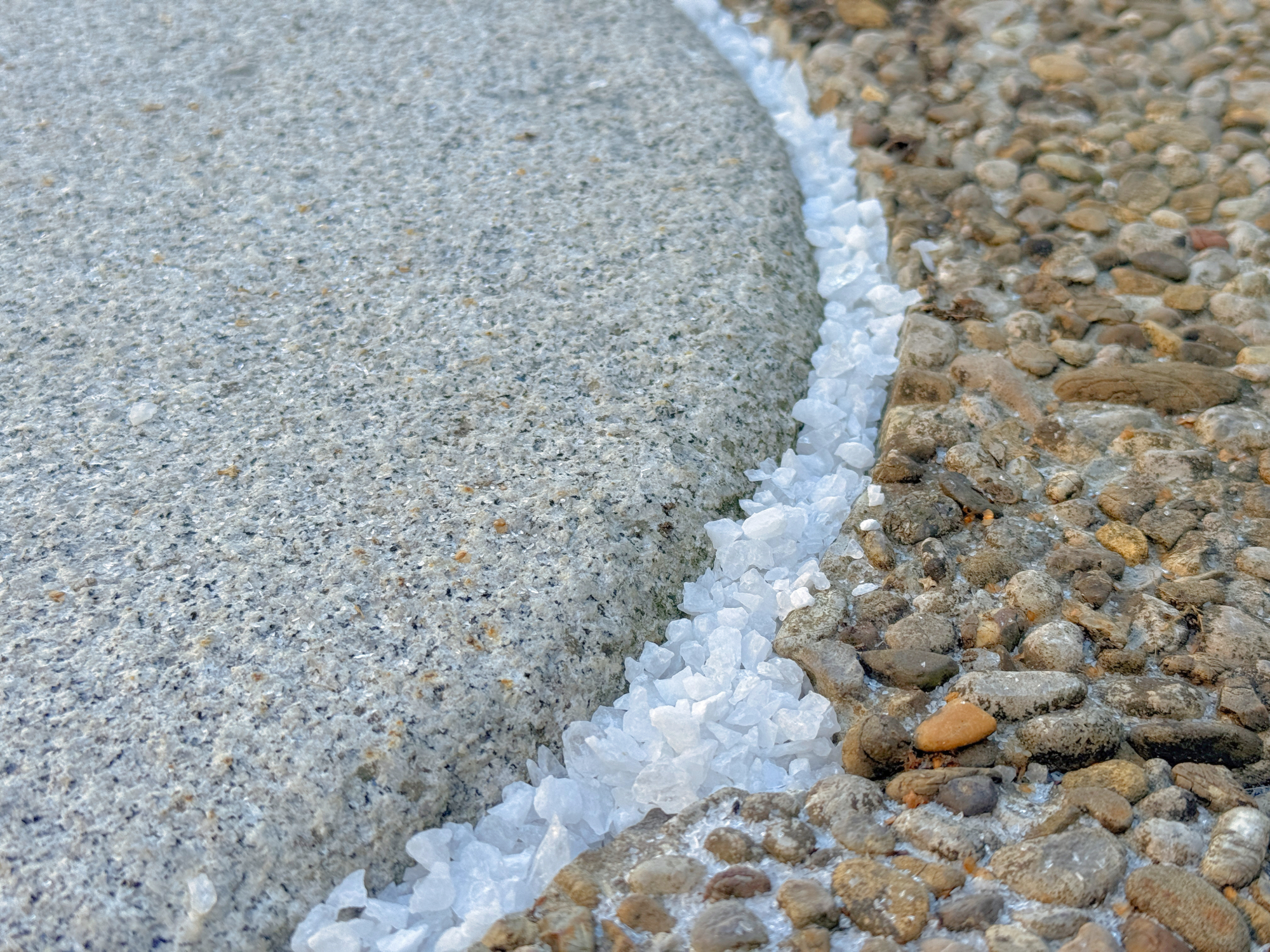 Weißes Bergkristall‑Granulat in Beutel auf hellem Rundfeld, umgeben von grober Steinpflasterung.
