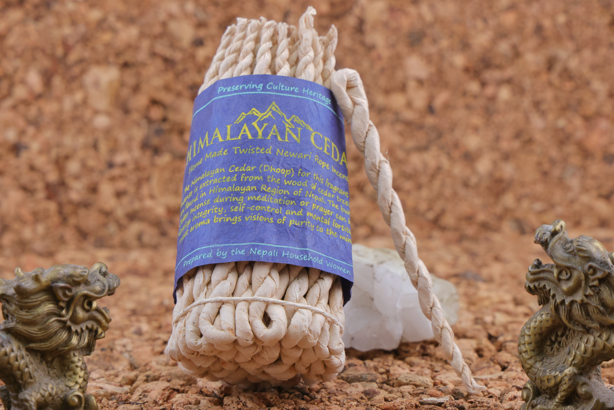 Handgedrehtes Himalaya‑Zeder‑Weihrauchseil mit blauem Etikett, dekoriert mit zwei bronzefarbenen Drachenfiguren und weißen Kristallen auf einer Korkunterlage.